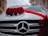 A white car is adorned with a decorative arrangement of vibrant red roses and white flowers on the hood. A red ribbon is elegantly draped across, enhancing the festive appearance. The emblem of the car manufacturer is prominently displayed on the front grille.