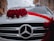 A white car is adorned with a decorative arrangement of vibrant red roses and white flowers on the hood. A red ribbon is elegantly draped across, enhancing the festive appearance. The emblem of the car manufacturer is prominently displayed on the front grille.