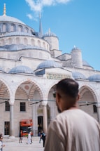 A large, ornate building with multiple domes and decorative arches, characteristic of Islamic architecture, dominates the scene under a clear blue sky. People are walking in front of the structure, and a man wearing a beige shirt is visible in the foreground, looking towards the building.