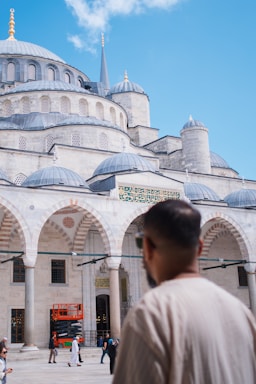 A large, ornate building with multiple domes and decorative arches, characteristic of Islamic architecture, dominates the scene under a clear blue sky. People are walking in front of the structure, and a man wearing a beige shirt is visible in the foreground, looking towards the building.