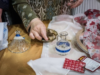 A person wearing a patterned garment handles small brown objects, possibly spices or herbs, using their fingers. The surface contains a decorative glass jar with a blue and white lace trim, a small pitcher, a red packet with writing, and folded textiles featuring floral designs.