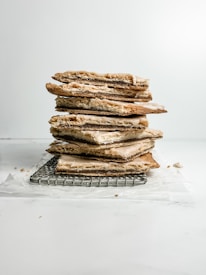 A stack of flaky, layered pastries is placed on a wire cooling rack. The pastries have light brown, crispy edges and a creamy filling that slightly oozes out. Crumbs and a piece of parchment paper are visible around the base, set against a simple white background.