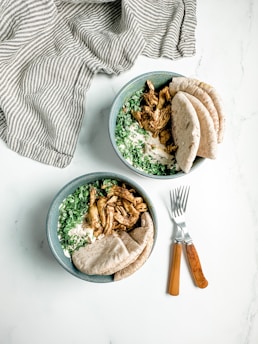 Two bowls are filled with sliced grilled chicken, chopped greens, creamy sauce, and served with pieces of pita bread. The bowls are accompanied by two wooden-handled forks on the side, placed on a white surface. A striped cloth is draped in the background.