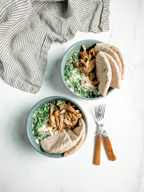 Two bowls are filled with sliced grilled chicken, chopped greens, creamy sauce, and served with pieces of pita bread. The bowls are accompanied by two wooden-handled forks on the side, placed on a white surface. A striped cloth is draped in the background.
