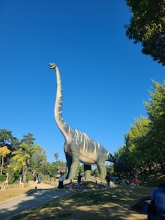 A large dinosaur sculpture stands in an outdoor area surrounded by trees and people. The dinosaur has a long neck and tail, resembling a sauropod, and features a light color with darker stripes. Several people are seen walking and observing the sculpture, and the clear blue sky enhances the vibrant setting.