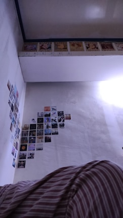The image shows a section of a room with several small posters or pictures arranged on the wall. The upper part of the wall features a row of 'WANTED' posters, reminiscent of those from anime or manga, while the lower part is filled with a diverse collection of smaller, colorful images. A portion of a ceiling with a reflective surface is visible, along with a bed covered in striped fabric in the foreground.
