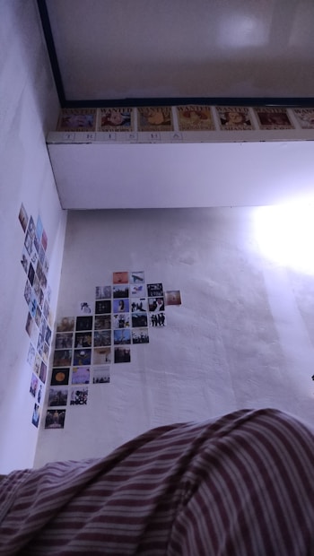 The image shows a section of a room with several small posters or pictures arranged on the wall. The upper part of the wall features a row of 'WANTED' posters, reminiscent of those from anime or manga, while the lower part is filled with a diverse collection of smaller, colorful images. A portion of a ceiling with a reflective surface is visible, along with a bed covered in striped fabric in the foreground.