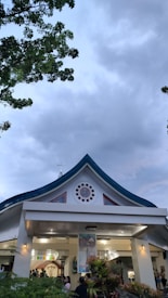 A large building with a distinct triangular roof is surrounded by greenery. The facade features a decorative circular design with red accents near the roof's peak. Various people are congregating at the entrance, which is well-lit and adorned with religious iconography. The cloudy sky and trees frame the view above.