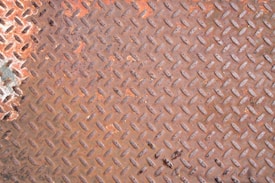 A close-up view of a rusted metal surface with a diamond plate pattern. The metal appears worn and oxidized, displaying various shades of orange and brown.