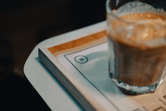A glass with a creamy, frothy beverage sits on a wooden tray. The tray appears atop a laminated piece of paper with an equal sign printed on it. The background is dark and out of focus.