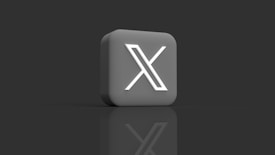 A 3D block with a prominent white 'X' symbol on its front surface, set against a dark background. The block appears to be glossy and has a reflective surface beneath it.