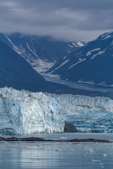 A massive glacier stretches across the scene, with intricate patterns of blue and white ice. The glacier leads into a serene body of water, reflecting the muted tones of the ice. Majestic, snow-capped mountains rise in the background under a blanket of cloudy sky.