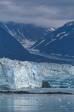 A massive glacier stretches across the scene, with intricate patterns of blue and white ice. The glacier leads into a serene body of water, reflecting the muted tones of the ice. Majestic, snow-capped mountains rise in the background under a blanket of cloudy sky.