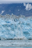 A massive glacier with light blue ice and jagged edges dominates the scene. The glacier is calving, with chunks of ice breaking off and splashing into the greyish water below. In the background, steep mountain slopes are partially visible under a heavy cloud cover.