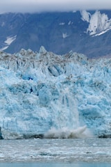 A massive glacier with light blue ice and jagged edges dominates the scene. The glacier is calving, with chunks of ice breaking off and splashing into the greyish water below. In the background, steep mountain slopes are partially visible under a heavy cloud cover.