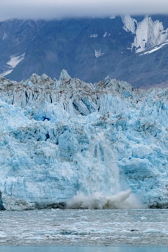 A massive glacier with light blue ice and jagged edges dominates the scene. The glacier is calving, with chunks of ice breaking off and splashing into the greyish water below. In the background, steep mountain slopes are partially visible under a heavy cloud cover.