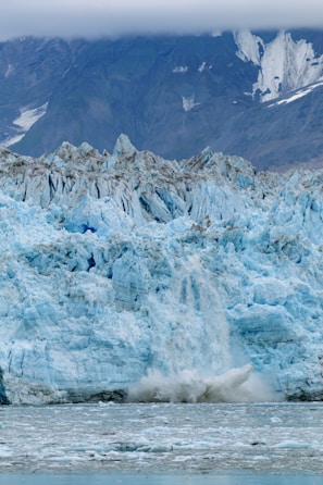 A massive glacier with light blue ice and jagged edges dominates the scene. The glacier is calving, with chunks of ice breaking off and splashing into the greyish water below. In the background, steep mountain slopes are partially visible under a heavy cloud cover.
