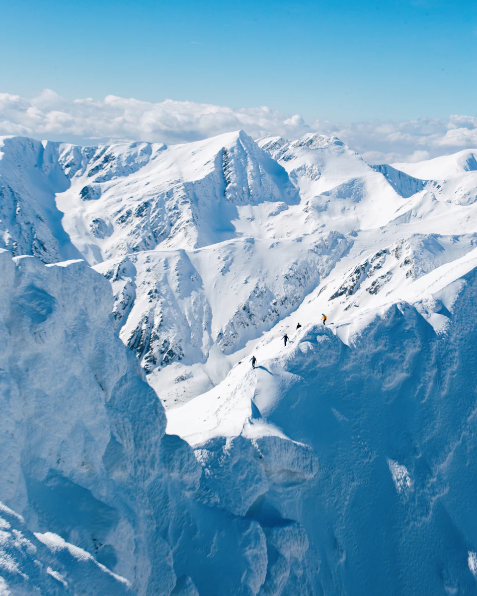 The image depicts a breathtaking view of snow-capped mountains under a clear blue sky. The mountain range is blanketed in pristine white snow, with several peaks reaching towards the heavens. A group of people can be seen hiking up one of the steep slopes, their figures small against the grandeur of nature.

The perspective of the image is from above, providing a bird's eye view of the majestic landscape below. The colors are predominantly blue and white, reflecting the wintry conditions on the mountainside.