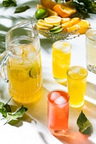 A sunlit scene features a glass pitcher of yellow lemonade with ice cubes and lime slices, surrounded by three tall glasses filled with different colored drinks. A plate with assorted citrus fruits, including lemons, limes, and oranges, is in the background. Bright light casts distinct shadows and highlights the vibrant colors.