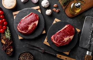 Two raw steaks are placed on black circular slate boards, surrounded by ingredients such as cherry tomatoes, garlic, onion, herbs, a bowl of salt, peppercorns, a bottle of olive oil, and dried mushrooms. A large meat cleaver rests nearby, all on a dark textured surface.