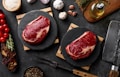 Two raw steaks are placed on black circular slate boards, surrounded by ingredients such as cherry tomatoes, garlic, onion, herbs, a bowl of salt, peppercorns, a bottle of olive oil, and dried mushrooms. A large meat cleaver rests nearby, all on a dark textured surface.