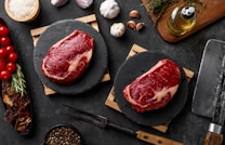 Two raw steaks are placed on black circular slate boards, surrounded by ingredients such as cherry tomatoes, garlic, onion, herbs, a bowl of salt, peppercorns, a bottle of olive oil, and dried mushrooms. A large meat cleaver rests nearby, all on a dark textured surface.