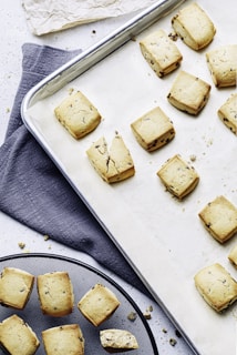 Several square-shaped cookies with chocolate chips are arranged on a baking sheet lined with parchment paper. Some cookies have visible cracks, and one has broken into pieces. The baking sheet is placed on a dark grey cloth with part of a cooling rack visible, which holds a few more cookies. Crumbs are scattered on the table surface, adding a rustic touch.
