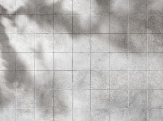 A grid of gray tiles with a slightly worn and textured surface. Shadows cast across the tiles give a sense of depth and varying light.