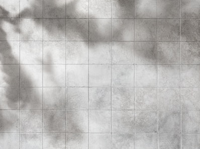 A grid of gray tiles with a slightly worn and textured surface. Shadows cast across the tiles give a sense of depth and varying light.