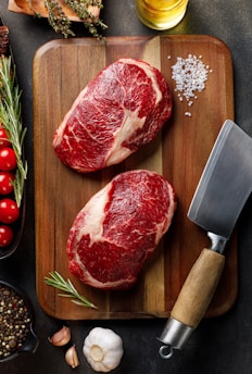 Two raw steaks are placed on a wooden cutting board, accompanied by a large cleaver on the side. Fresh herbs such as rosemary, thyme, and ingredients like cherry tomatoes, garlic cloves, and peppercorns are arranged around the board. A sprinkle of coarse salt and a small bottle of oil complete the setting.
