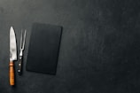 A large kitchen knife with a wooden handle and a meat fork with a black handle are placed next to a black rectangular cutting board on a textured dark surface.