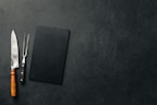 A large kitchen knife with a wooden handle and a meat fork with a black handle are placed next to a black rectangular cutting board on a textured dark surface.