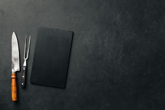 A large kitchen knife with a wooden handle and a meat fork with a black handle are placed next to a black rectangular cutting board on a textured dark surface.
