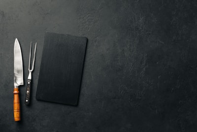 A large kitchen knife with a wooden handle and a meat fork with a black handle are placed next to a black rectangular cutting board on a textured dark surface.
