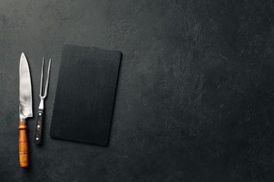 A large kitchen knife with a wooden handle and a meat fork with a black handle are placed next to a black rectangular cutting board on a textured dark surface.