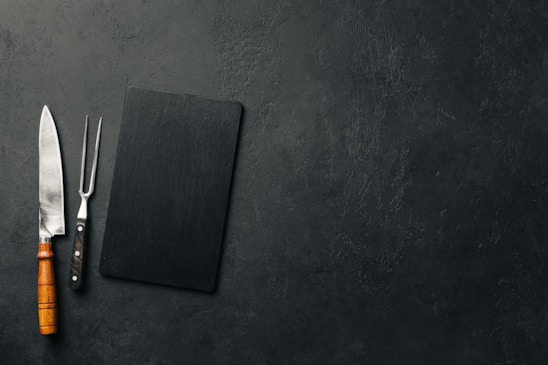 A large kitchen knife with a wooden handle and a meat fork with a black handle are placed next to a black rectangular cutting board on a textured dark surface.