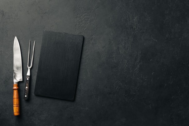 A large kitchen knife with a wooden handle and a meat fork with a black handle are placed next to a black rectangular cutting board on a textured dark surface.
