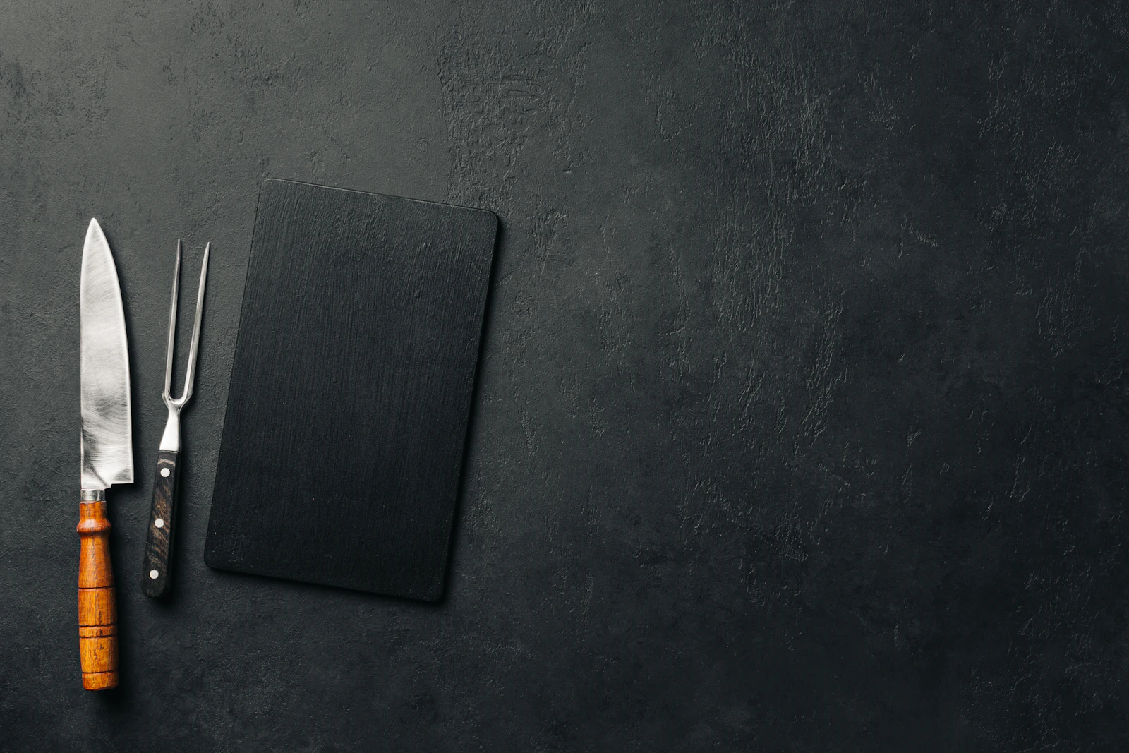 Top-down chef's knife, carving fork and black slate board on dark stone