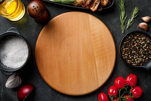 A wooden circular cutting board is surrounded by various cooking ingredients. On the left, there is a bowl of coarse salt and a peeled red onion half. At the top, a bottle of olive oil and a wooden pepper grinder sit beside rosemary sprigs and sun-dried tomatoes. On the right, a bowl of whole peppercorns is accompanied by more rosemary and garlic cloves. Below are vine-ripened cherry tomatoes.