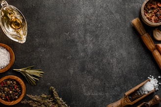 A dark textured surface serves as the backdrop for an array of culinary items, including bowls containing salt, peppercorns, and dried chili peppers. A sprig of rosemary lies next to a small carafe of oil, while a wooden brush and a garlic clove add to the rustic cooking theme. Wooden scoops hold coarse salt, complementing the organic and earthy feel.