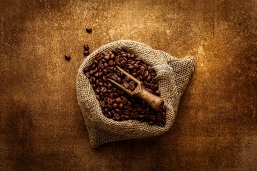 A burlap sack filled with roasted coffee beans is placed on a textured brown surface. A small wooden scoop rests on top of the beans, suggesting a rustic and natural setting.