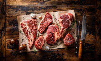 A rustic wooden table displaying a variety of beautifully marbled prime beef cuts with fresh herbs and butcher tools nearby.