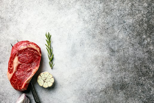 A raw cut of marbled meat is placed on a textured surface alongside a sprig of rosemary and a halved bulb of garlic. The surface has a speckled gray appearance that provides a rustic backdrop.