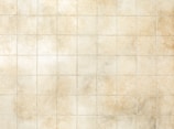 A grid of uniform, square tiles in a soft beige color with subtle texture. Some tiles have variations in shading, giving a weathered, rustic appearance.