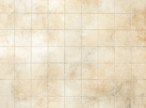 A grid of uniform, square tiles in a soft beige color with subtle texture. Some tiles have variations in shading, giving a weathered, rustic appearance.