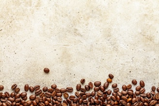 A collection of coffee beans is scattered across the bottom of a textured, light brown surface. The arrangement creates an organic border, while the beans are various shades of brown and appear glossy.