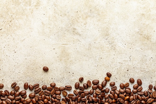 A collection of coffee beans is scattered across the bottom of a textured, light brown surface. The arrangement creates an organic border, while the beans are various shades of brown and appear glossy.