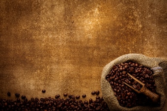 A burlap sack filled with dark coffee beans is placed on a textured brown surface. Some beans are scattered across the surface, with a wooden scoop resting on top of the beans. The overall composition has a rustic and earthy feel.