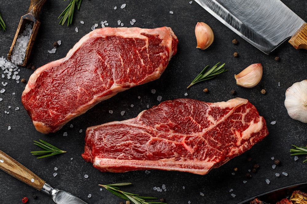 two steaks on a cutting board next to garlic, garlic, and a knife