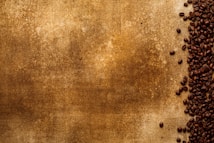 A textured brown background covers most of the image with a collection of dark roasted coffee beans scattered along the right side. The beans are densely packed, creating a contrast between the smooth texture of the beans and the rougher, mottled surface of the background.